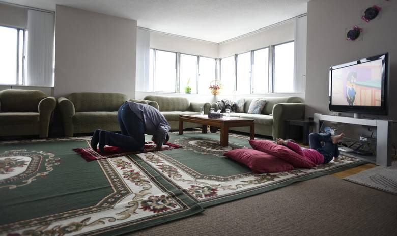 Unable to go to his mosque for prayer service, Ahmad Al Rasoul prays in his family’s apartment while daughter Sava, 3, watches TV.