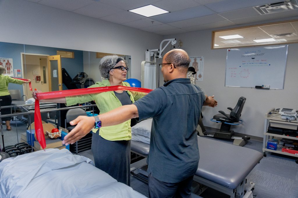 A woman stands with her arms outstretched using resistance bands while a physiotherapist faces her and mirrors her pose