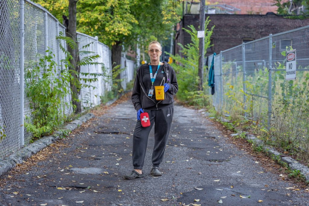 A person stands outside in an alleyway lined with chain link fences. They are wearing gloves and are holding a container for sharps