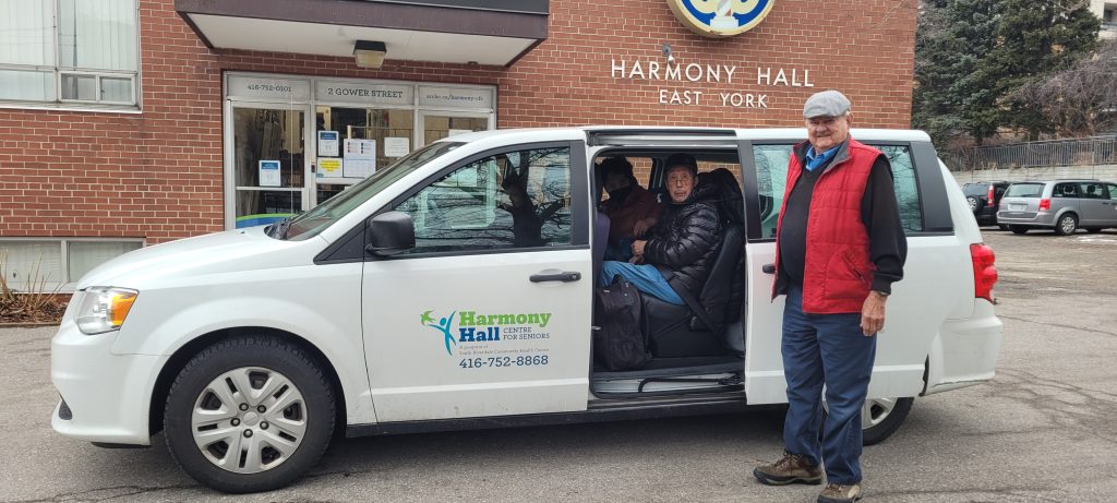 A person sits inside of a van with an open door. Next to the open door stands a Harmony Hall driver