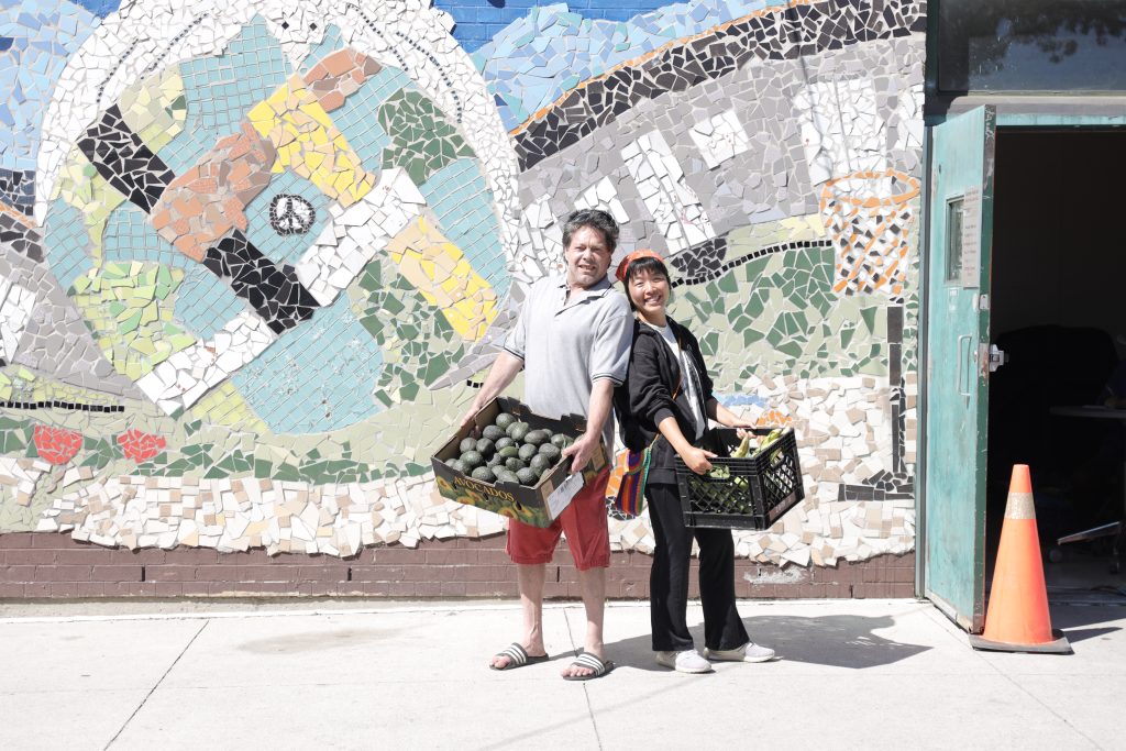 Two people stand outside in front of a mosaic wall. Each are holding a bin full of fresh produce