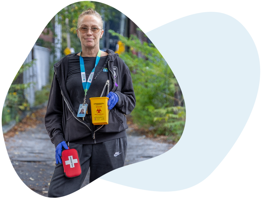 A person stands outside in an alleyway lined with chain link fences. They are wearing gloves and are holding a container for sharps