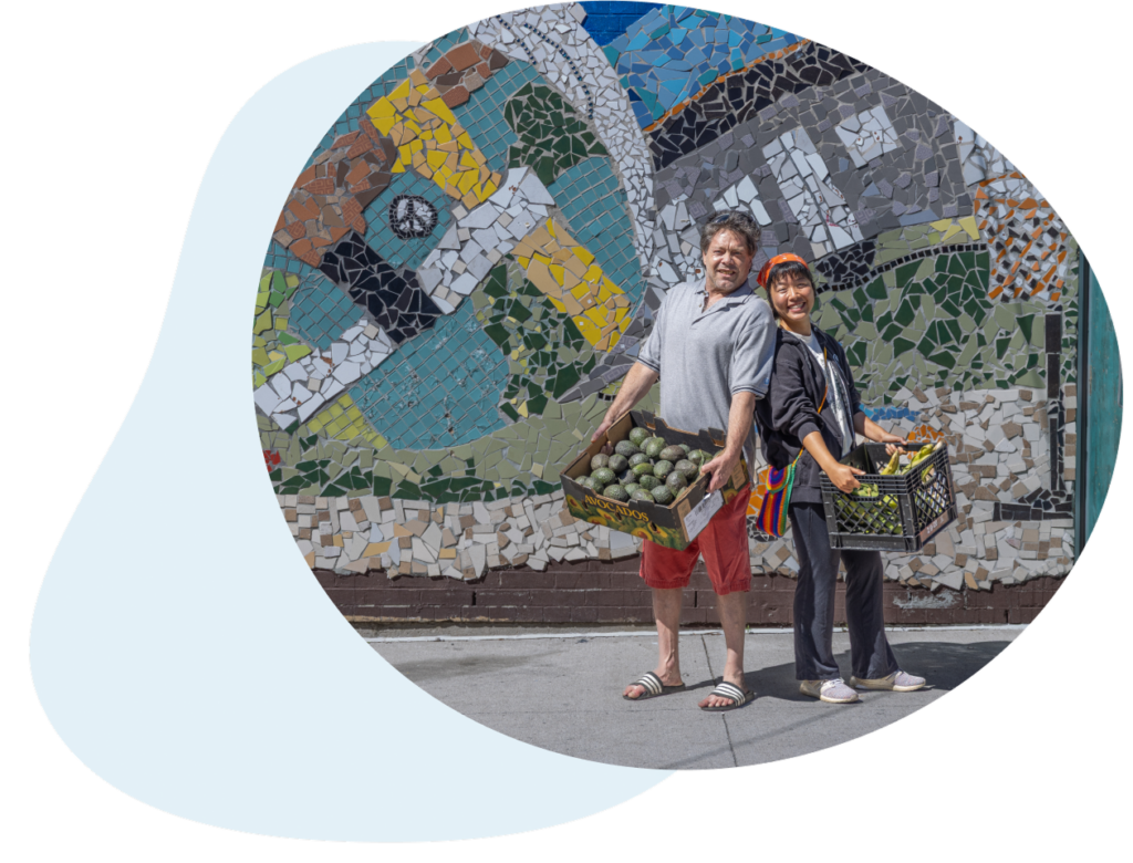 Two people stand outside in front of a mosaic wall. Each are holding a bin full of fresh produce